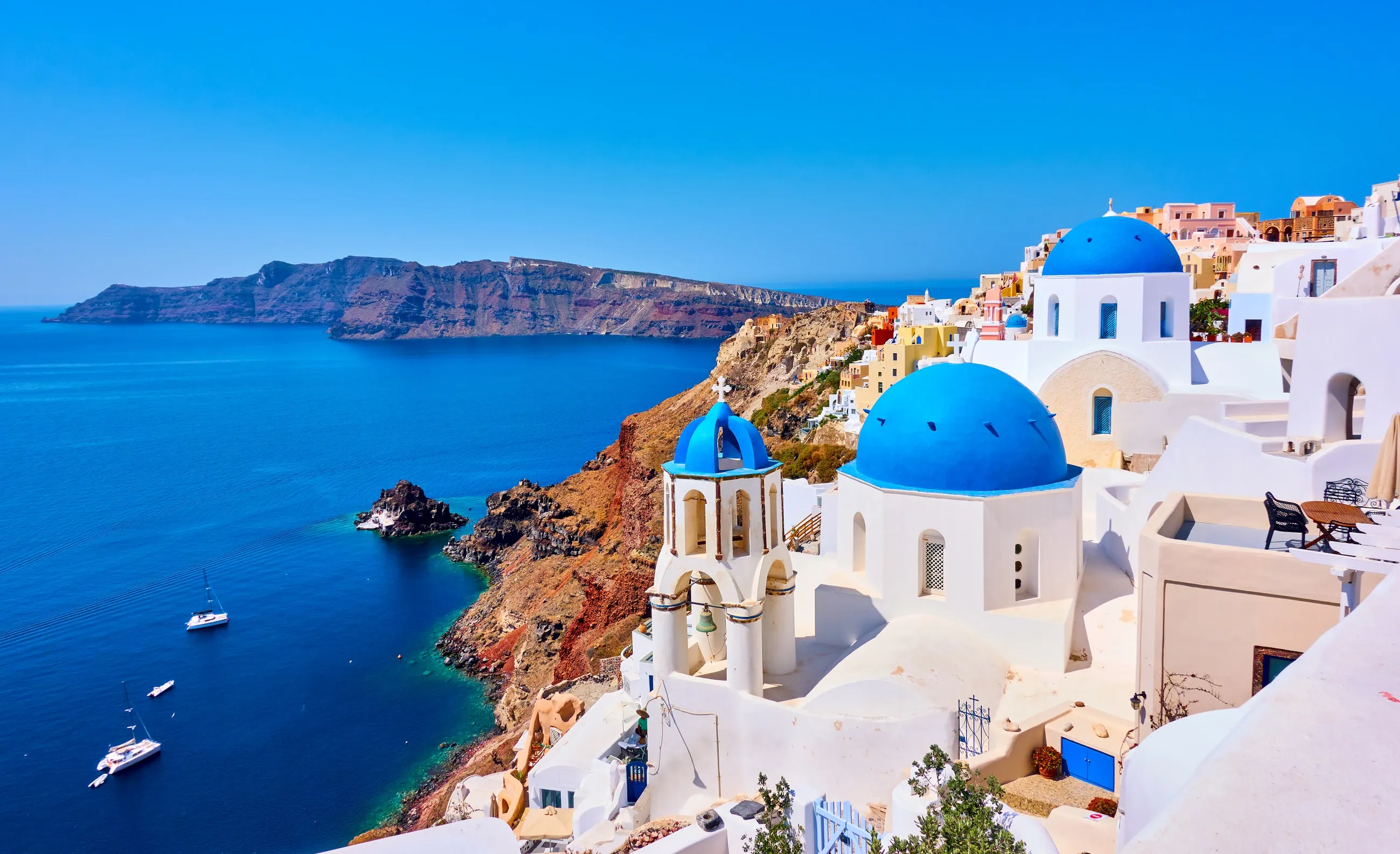 Vita byggnader med blå kupoler på en klippa vid det djupblå Egeiska havet på Santorini, Grekland, med segelbåtar i viken